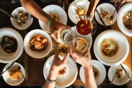 shot from above overlooking a table top of food with four people's arms outstretched touching their drinks glasses together in cheers