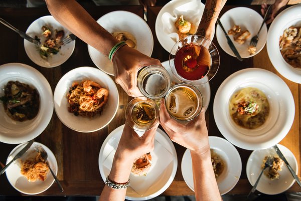 shot from above overlooking a table top of food with four people's arms outstretched touching their drinks glasses together in cheers