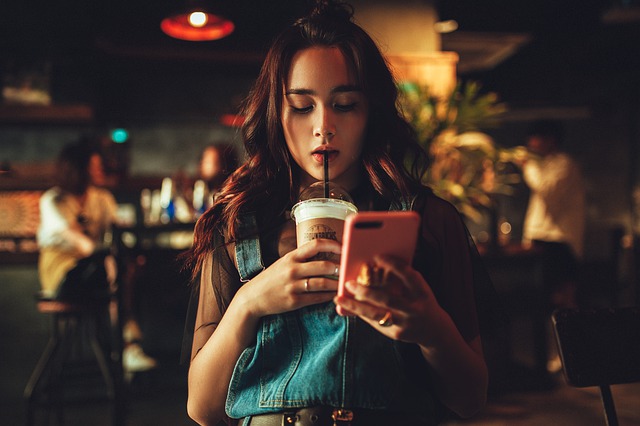 teenage girl holding an iced drink if one hand and a phone in the other. she is looking at the screen fully absorbed in it and not noticing anything else. She is in need of a digital detox.