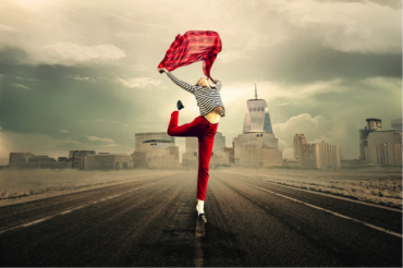 female wearing red troudrs and a stripy long sleveed top, jumping in the air while holding a rd scarf above her head. the background is a cityscape and it is meant to represent her lifestyle choices