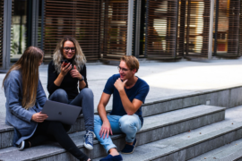 group of three college students sat on outsde steps at fron tof college building. They are laughing together and looking as though they are stress free