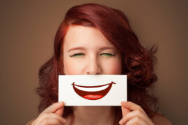 Face of an auburn haired woman holdng a piece of card over her mouth with apicture of a cartoon smile mouth on it