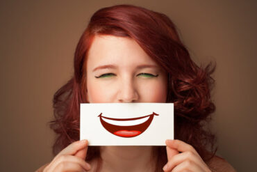 Face of an auburn haired woman holdng a piece of card over her mouth with apicture of a cartoon smile mouth on it