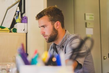 man looking stressed sat at his desk surrounded by stationery looking as though he is suffering from chronic pain through workiing at his office job