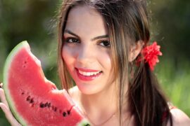 close up of a young brunette female face. She has her hair tied back in a side ponytail fastened with a red flower band and she is holding a slice of watermelon