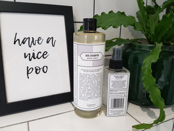 A bottle of Norfolk Natural Living Dog Shampoo and Dog Cologne on a white metro tiled shelf next to a black framed picture that says have a nice poo and a green plant in a green potis on the other side