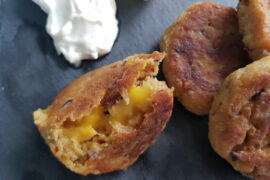 Close up of 4 vegan cheese falafels on a grey slate board with lemon slices and a dollopof aioli inthe corner. One falafel has been halved revealing a melted vegan cheese centre