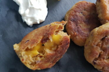 Close up of 4 vegan cheese falafels on a grey slate board with lemon slices and a dollopof aioli inthe corner. One falafel has been halved revealing a melted vegan cheese centre